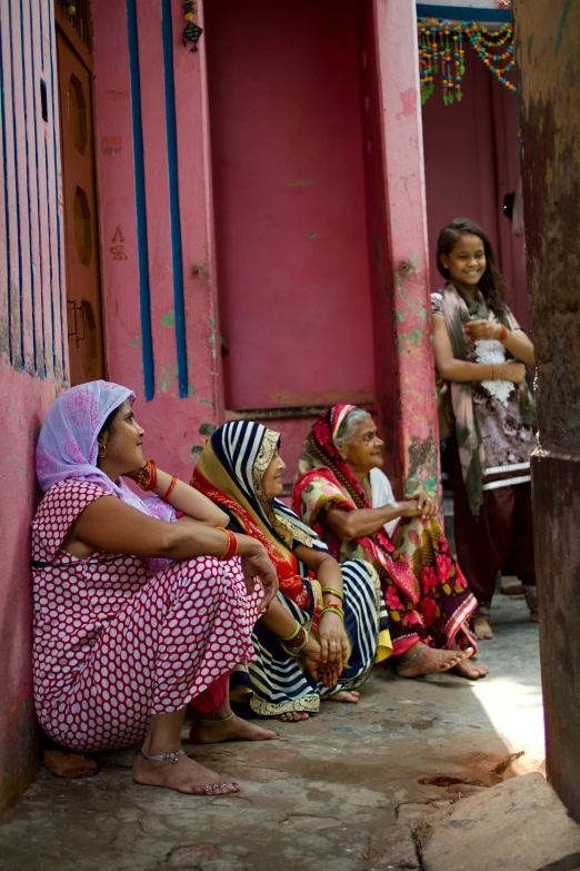 two women and one man in traditional dress sit together against a pink building