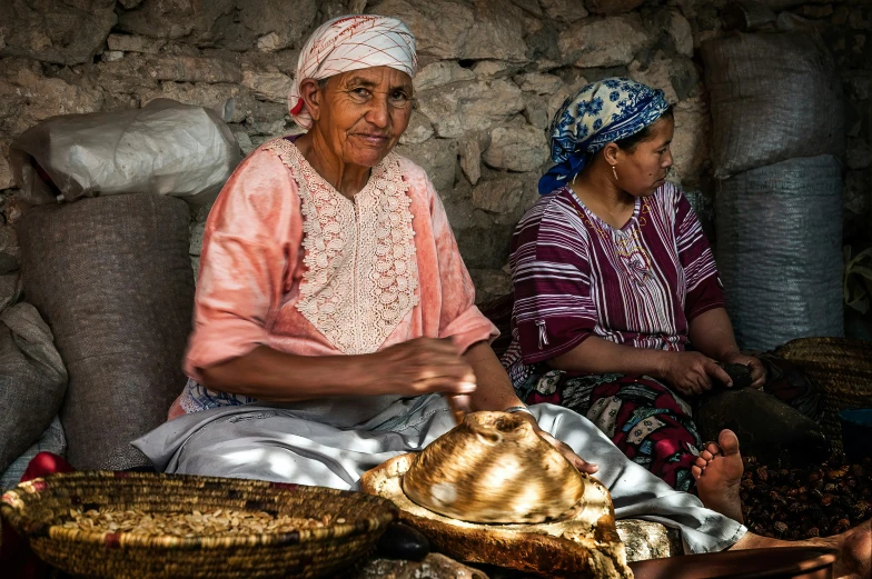 two women sit on a stone wall with baskets and corn