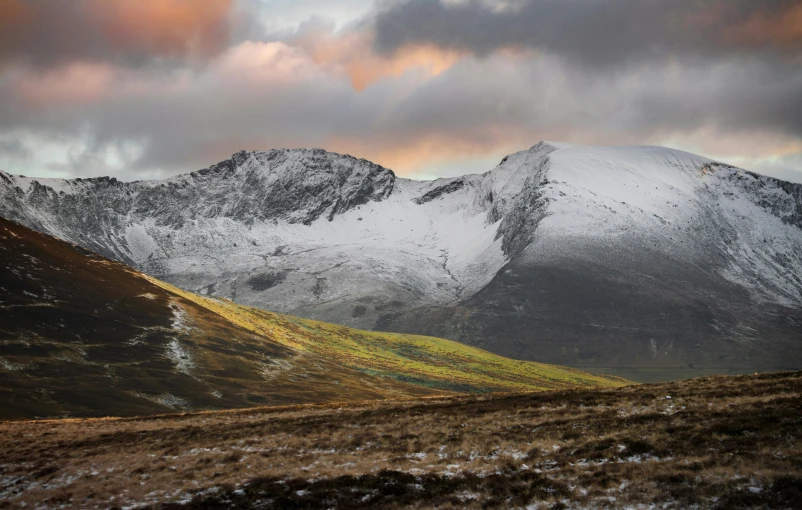 two mountains under a cloudy sky and a brown grass field