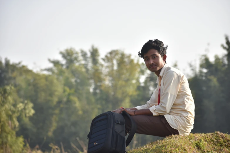 a man with long hair and a white shirt and brown pants sits on grass next to a black suitcase