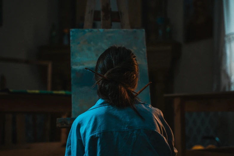 woman in blue shirt standing next to easel with painting brush in her hand