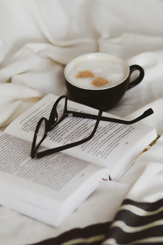 a coffee cup with glasses rests on an open book on a bed