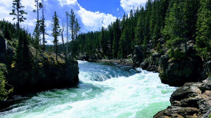 a waterfall running between mountains and forests on a sunny day