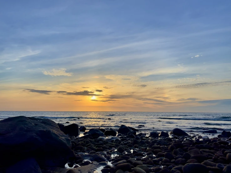 the sun is setting over the water on a rocky shore