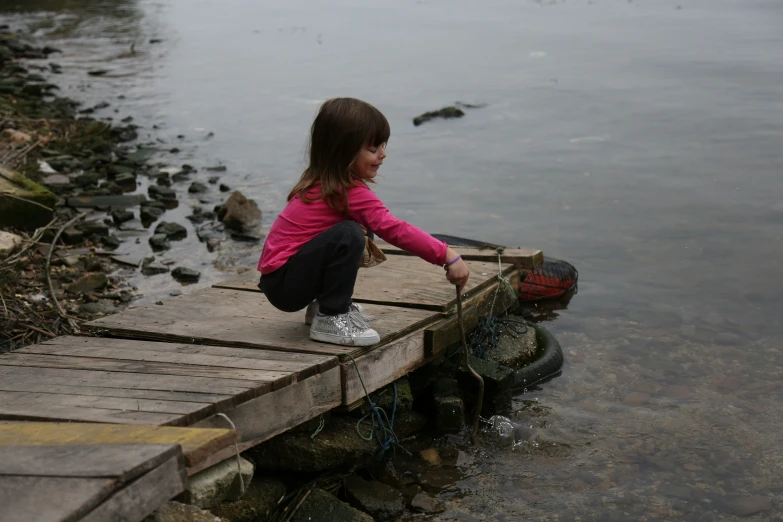 little girl with pink jacket and sneakers sitting on pier next to lake