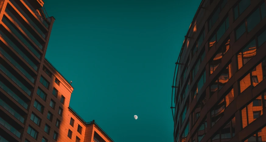 a half moon rises over the side of tall buildings