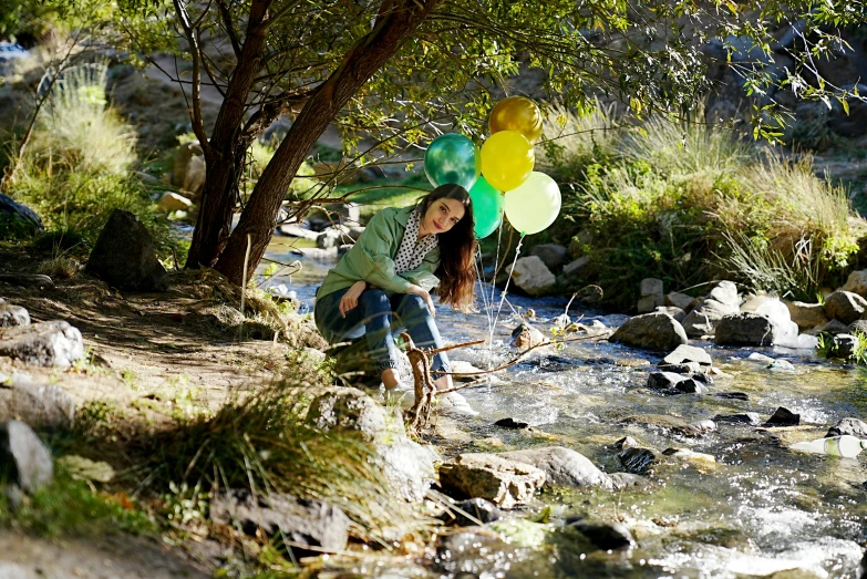 girl sitting on nch next to water and tree with balloons