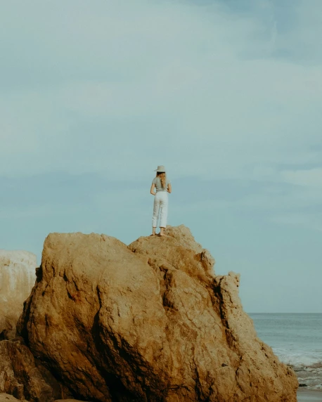 a woman in a white shirt and hat standing on a rock near the ocean