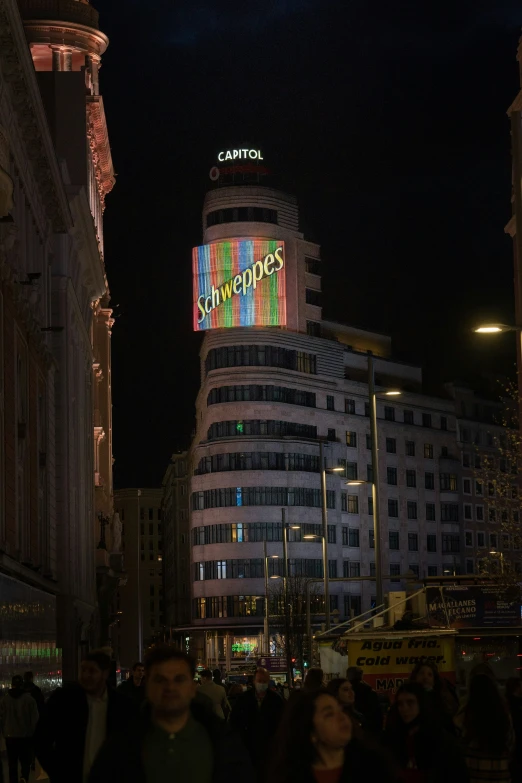 people walking on the street at night with a building lit up in bright colors