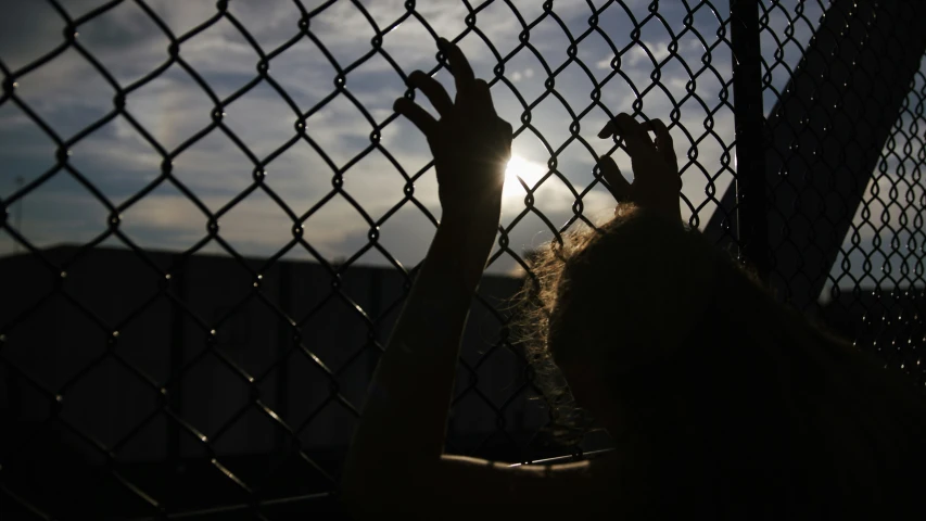 a person reaching up through a fence toward a setting sun