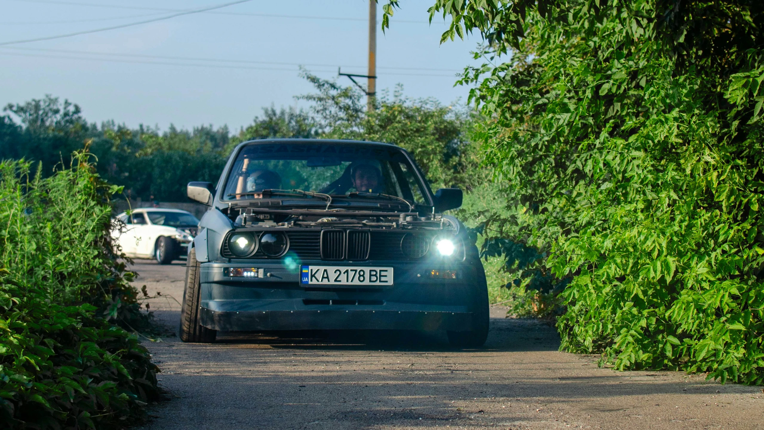 an old blue car with a hood up driving on a narrow road
