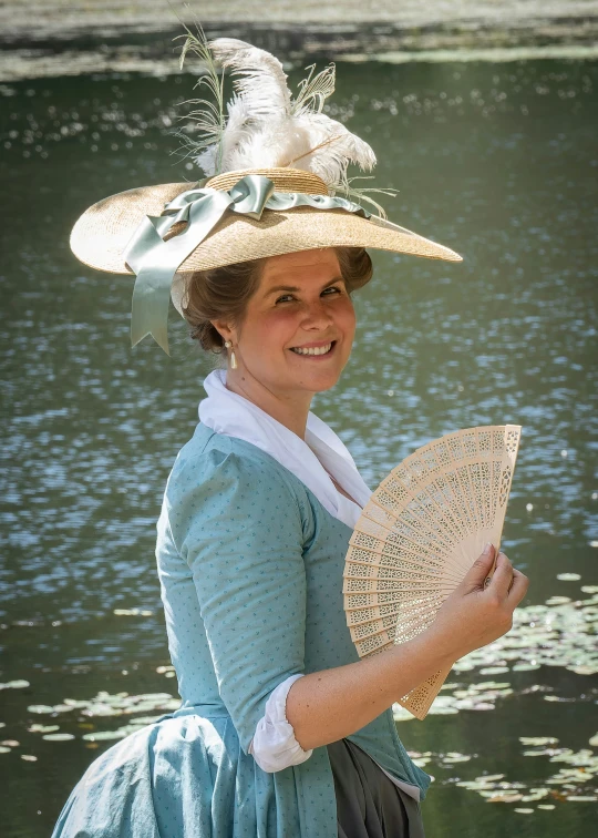 lady in a blue dress by a body of water holding a hat