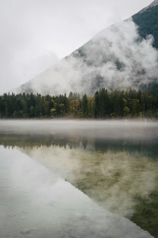 a foggy mountain is seen next to a still lake