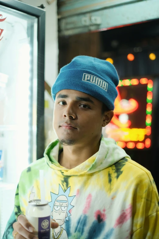 a guy in a blue hat and tie dyed shirt is holding a drink and standing by a vending machine