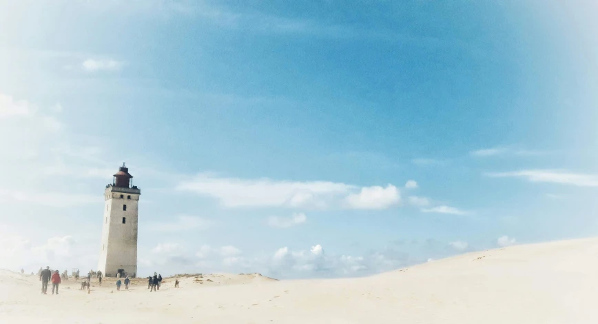 a group of people walking across a sand covered field near a light house