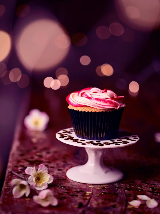 cupcake on pedestal next to flower petals with lights behind