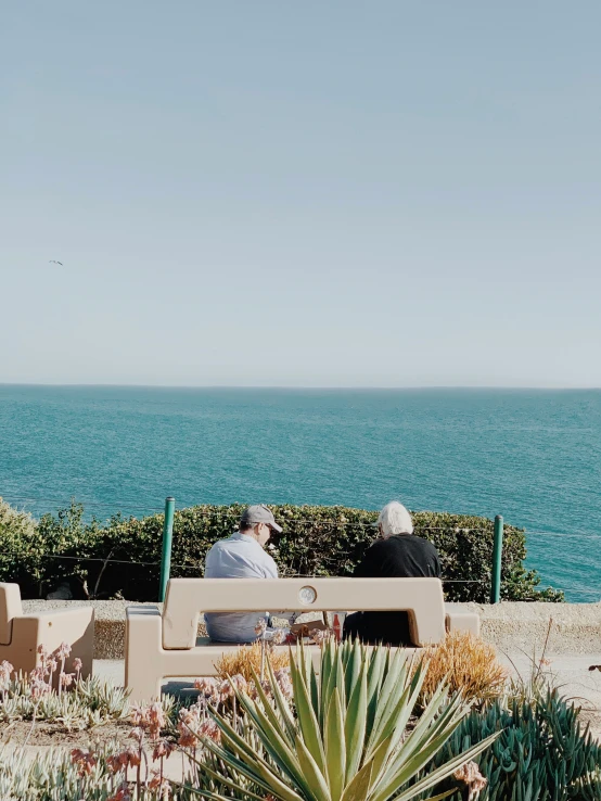 two elderly people sitting on a bench facing the ocean