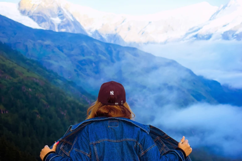 a woman wearing a hat and jeans stands in the mountains overlooking a mountain range