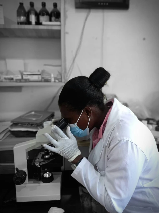 a woman with gloves, glasses and surgical mask looking into a microscope