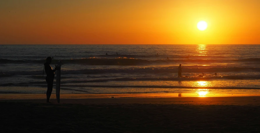 the man stands near his surfboard watching the sunset on a beach