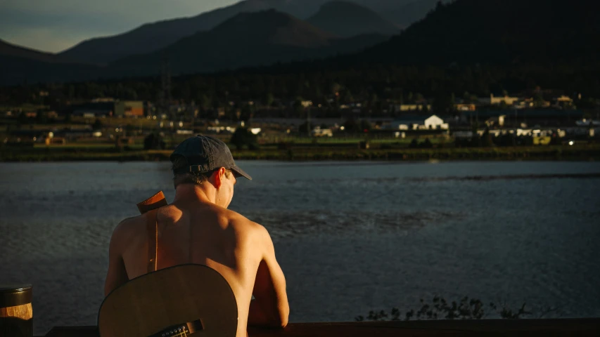 shirtless male playing guitar near water and hills