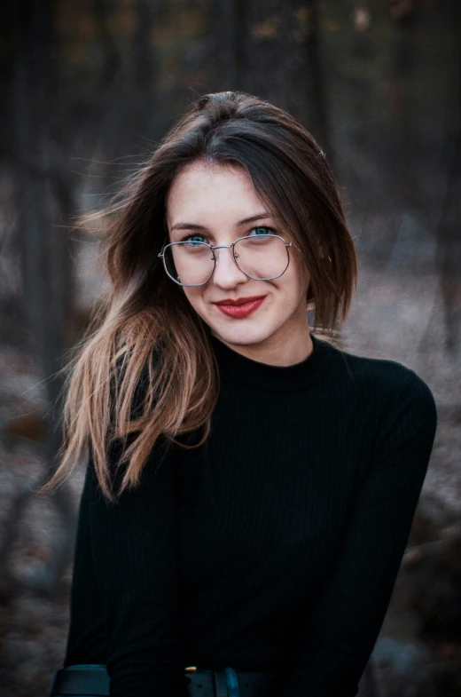 a woman with glasses and brown hair stands in a wooded area