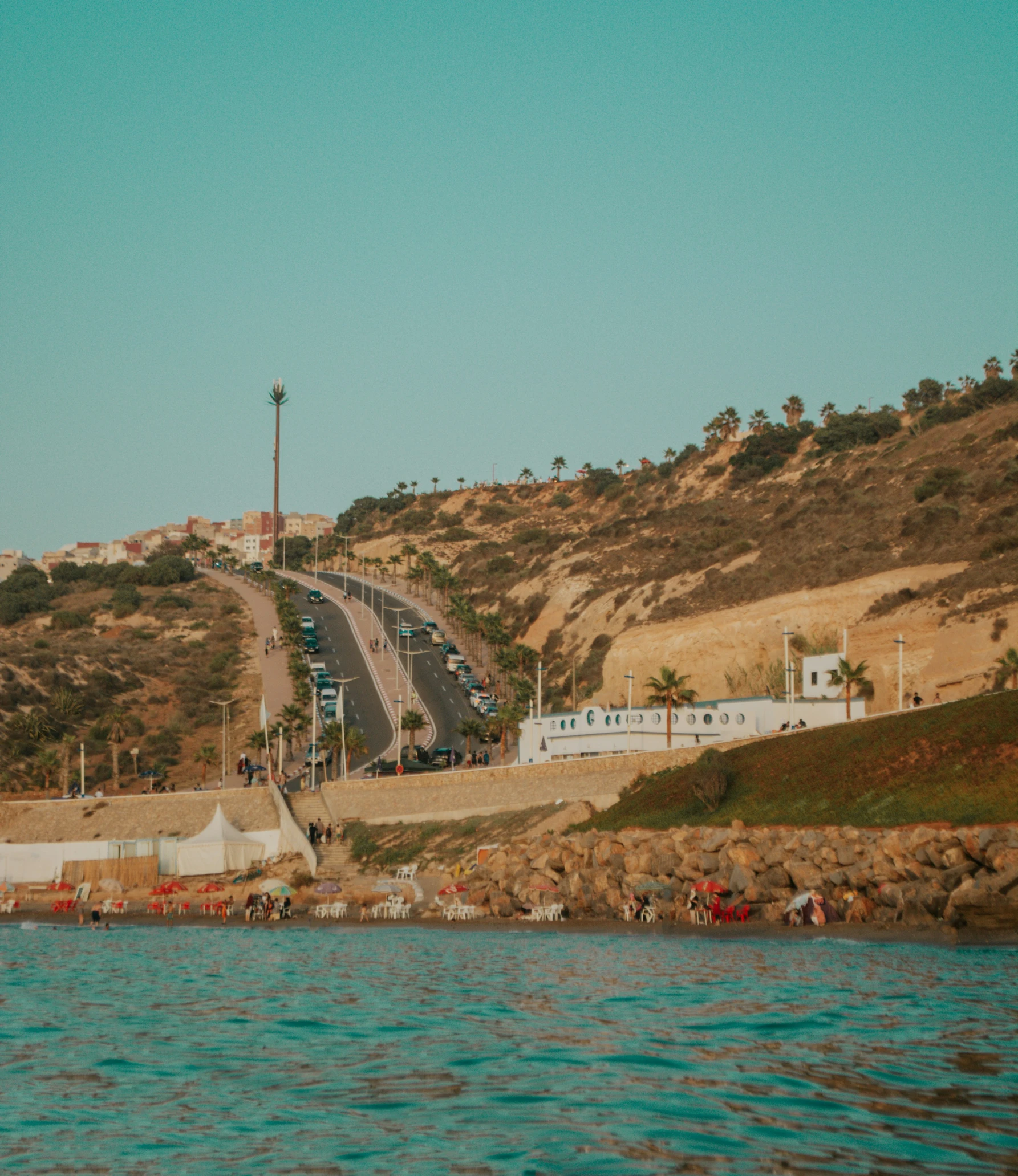 a group of cars driving on a road that's between the ocean and a beach