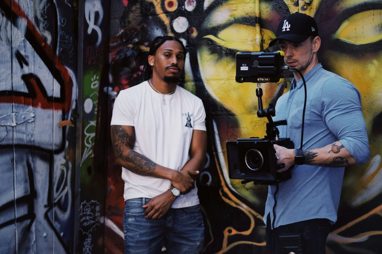 two men stand together in front of a wall with graffiti and a camera on it