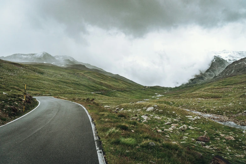 a road near a grassy hill and snow covered mountains