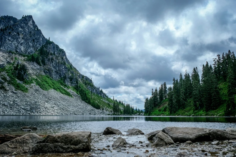 a cloudy, wooded area next to a lake with rocks and trees