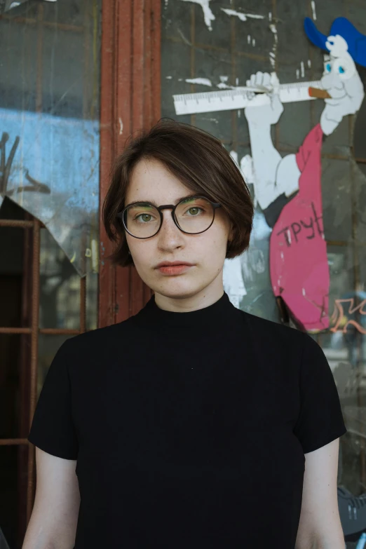 a woman with glasses stands near a graffiti covered wall