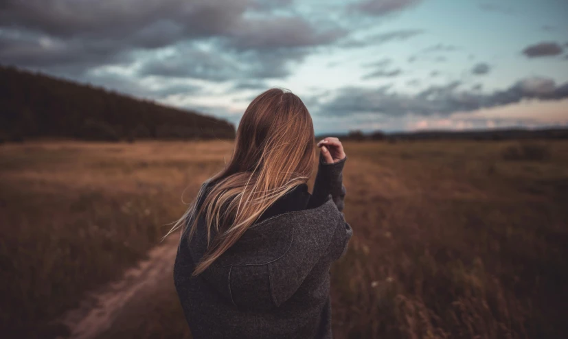 a woman stands in an open field, her long brown hair back, and looks over her shoulder