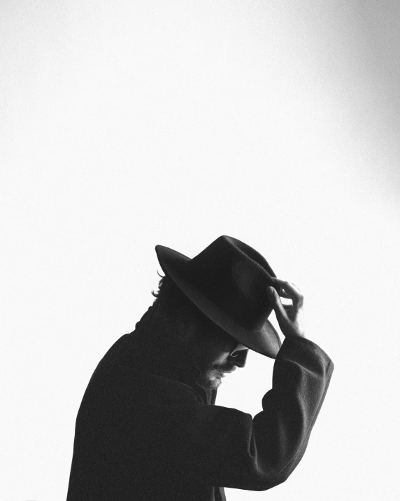 a man stands with his hands to his head and wears a fedora