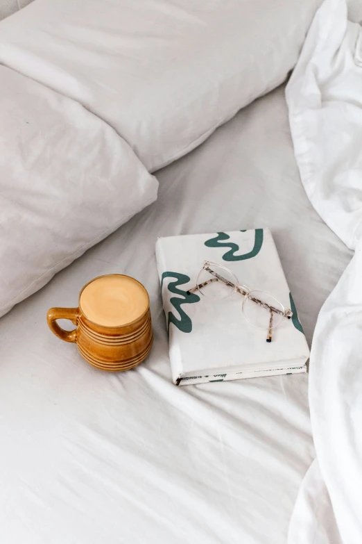 a cup and small tea pot laying on the bed with a white pillow and pillows