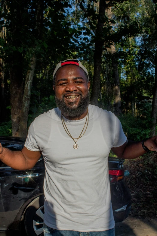 a man smiling, with his hands up with trees and cars behind him