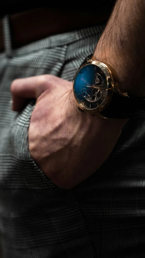 a male wearing a gold wrist watch with a blue strap