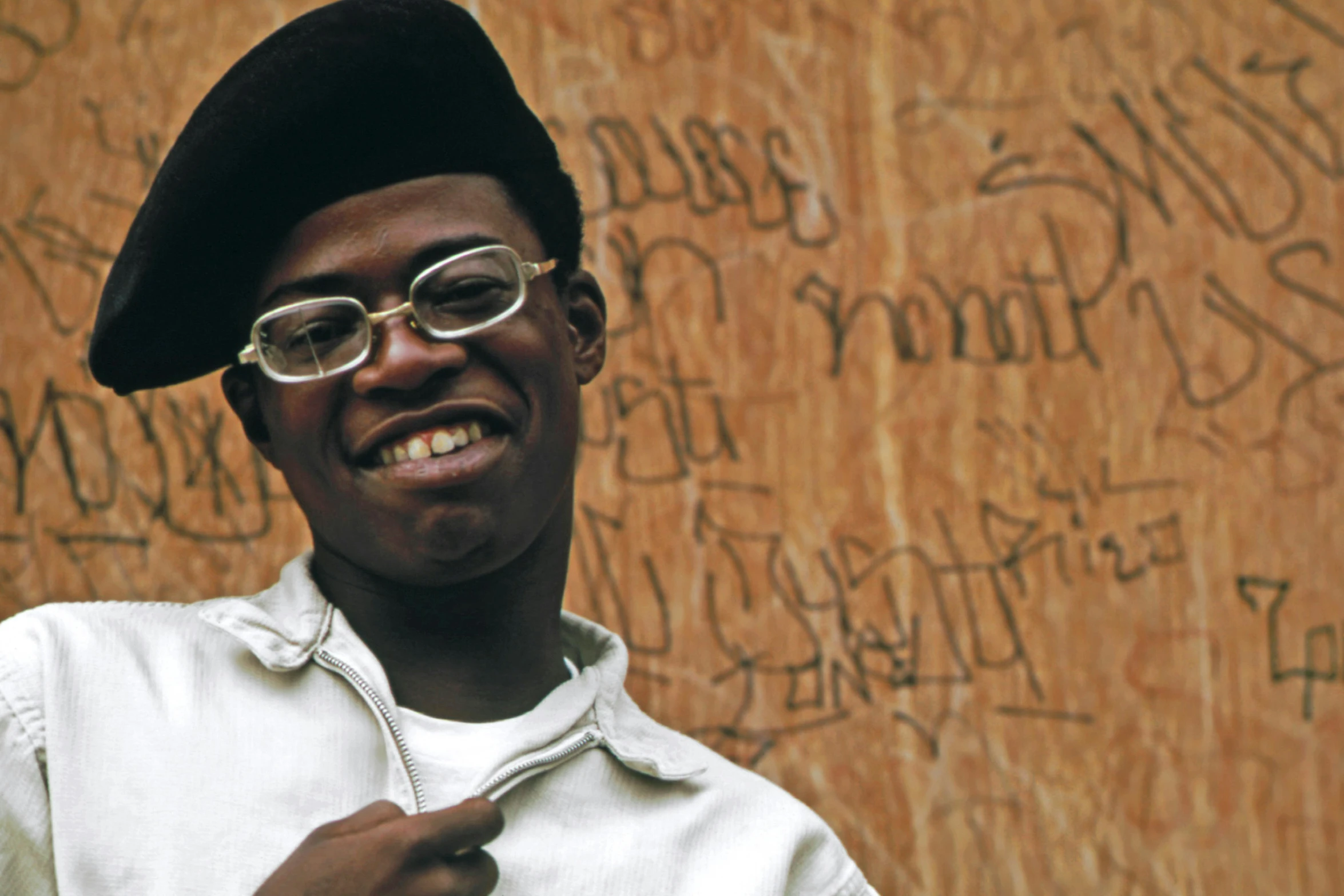 man wearing glasses and a hat by wall with writing on it