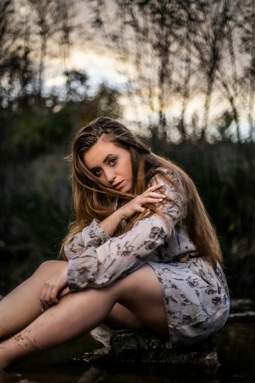 a woman with long hair sitting down on a river bank