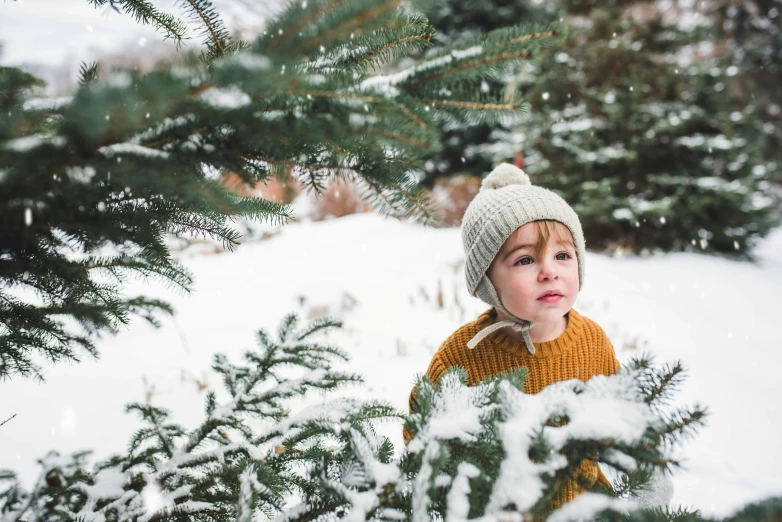 a small child in yellow sweater and knit cap with pine tree nches in the background