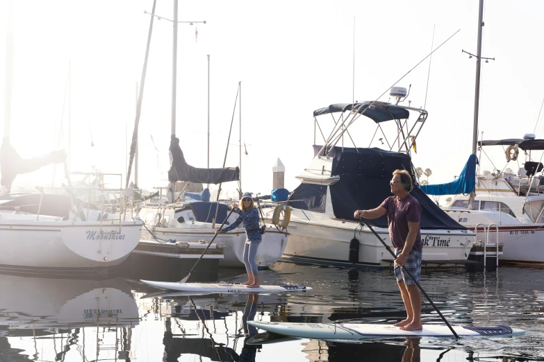 two people standing on surfboards and looking at the sailboats