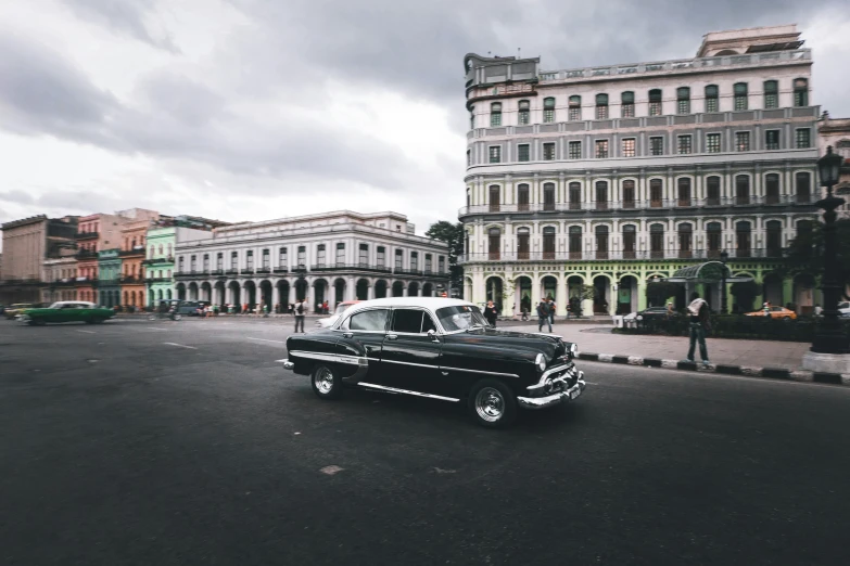 a classic car driving in the street with tall buildings in the background