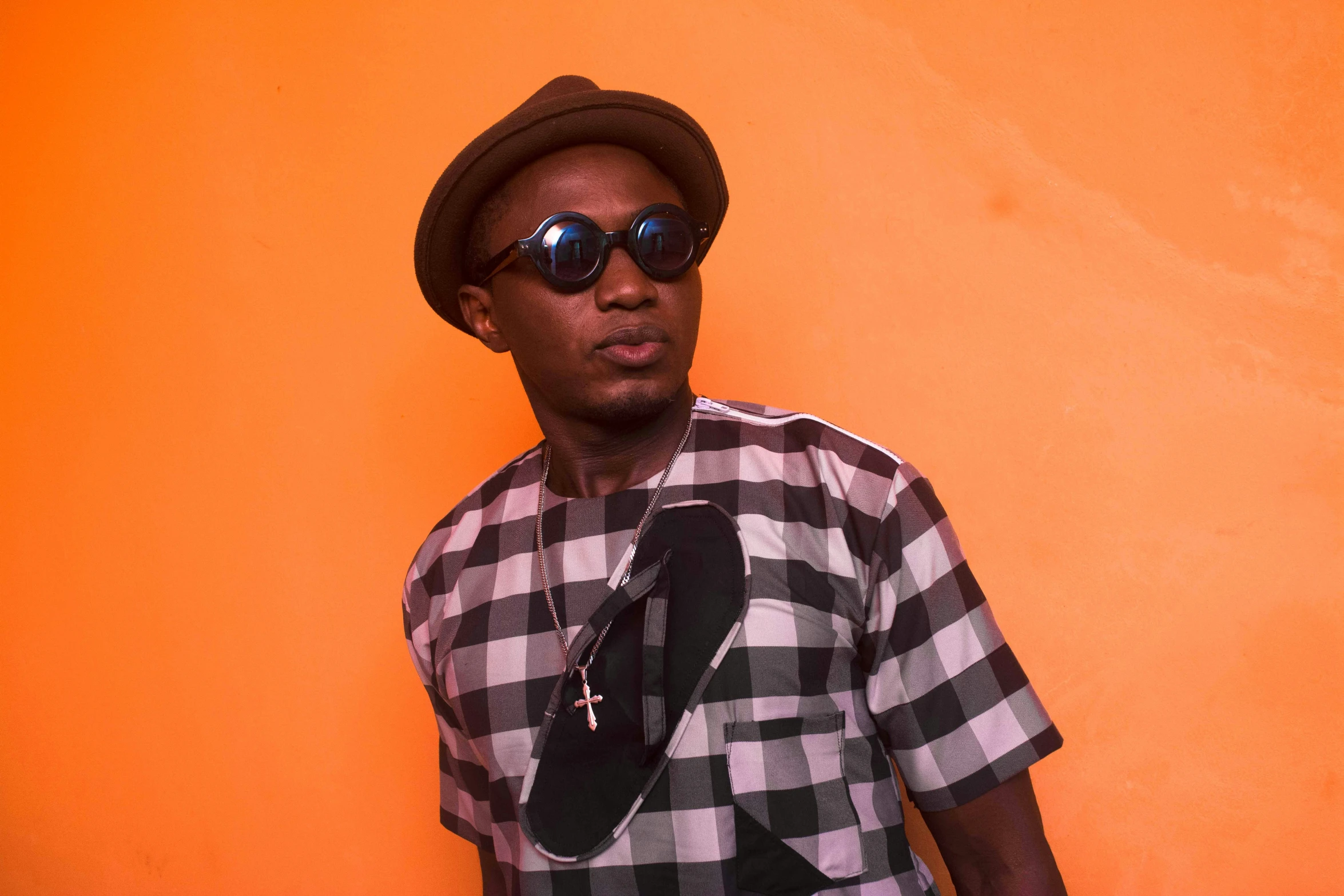 a man with dark sunglasses and a hat, standing against an orange wall