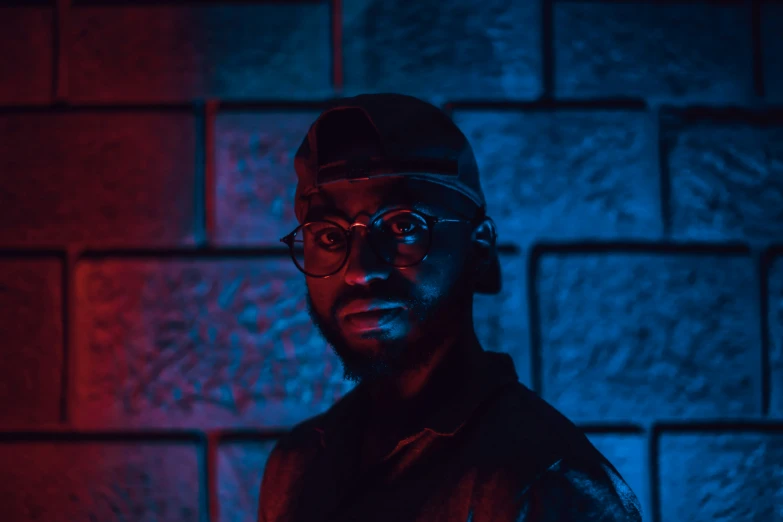 a man with glasses and a black hat in front of a brick wall