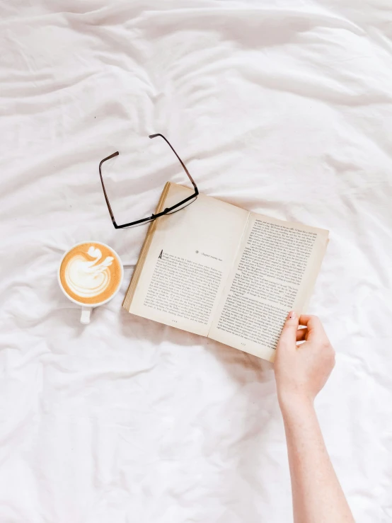 a book, glasses and a cappuccino are lying next to each other on a bed