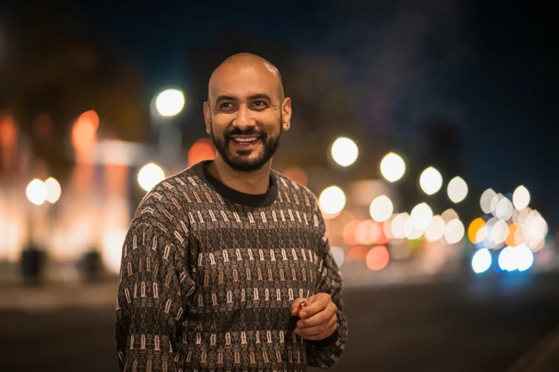 a smiling man wearing a patterned sweater stands near a dark street