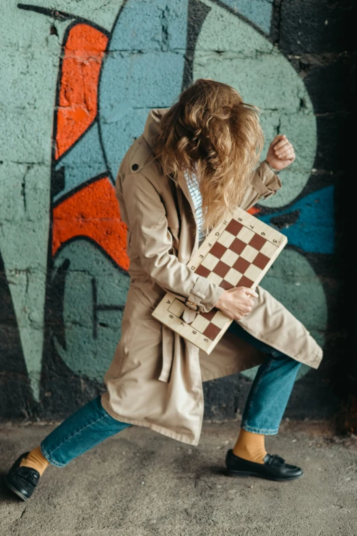 the woman is holding a cardboard checkerboard near a graffiti wall