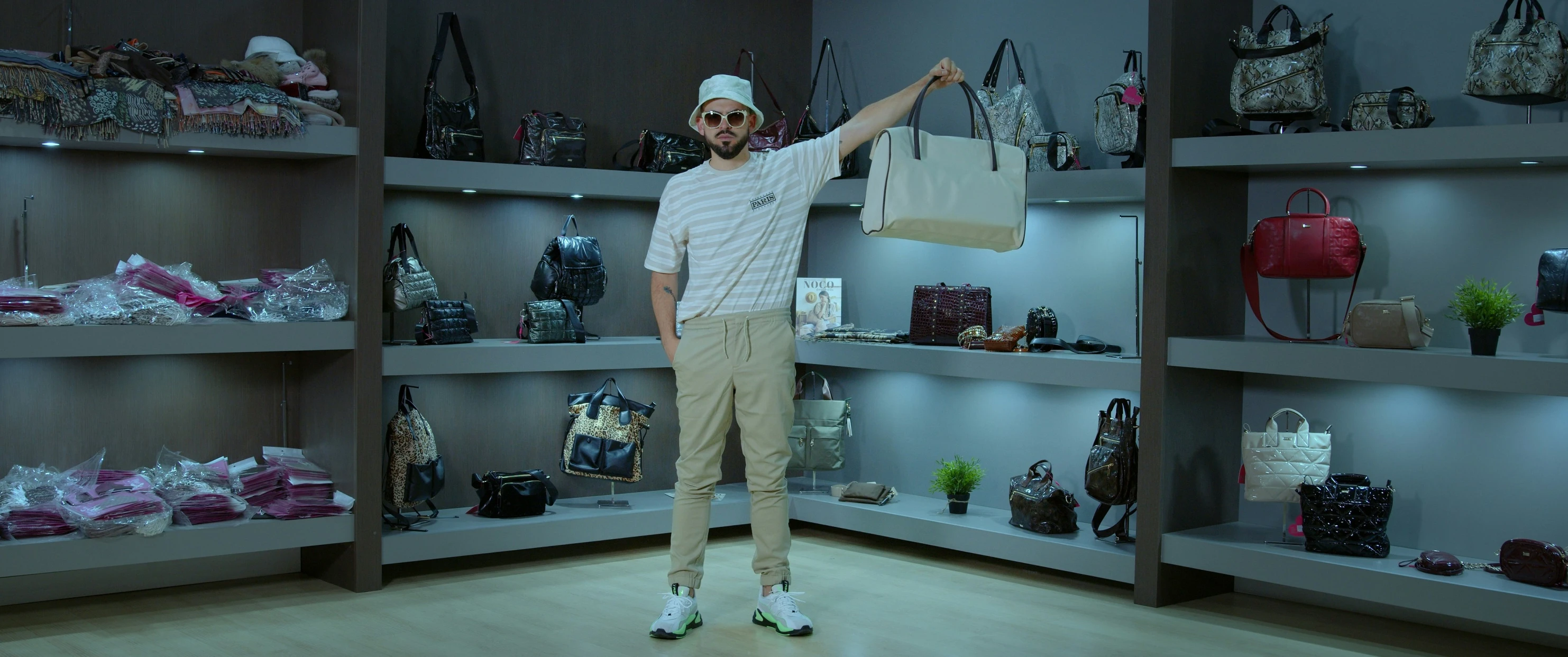 the man has his arms out as he stands next to shelves with handbags