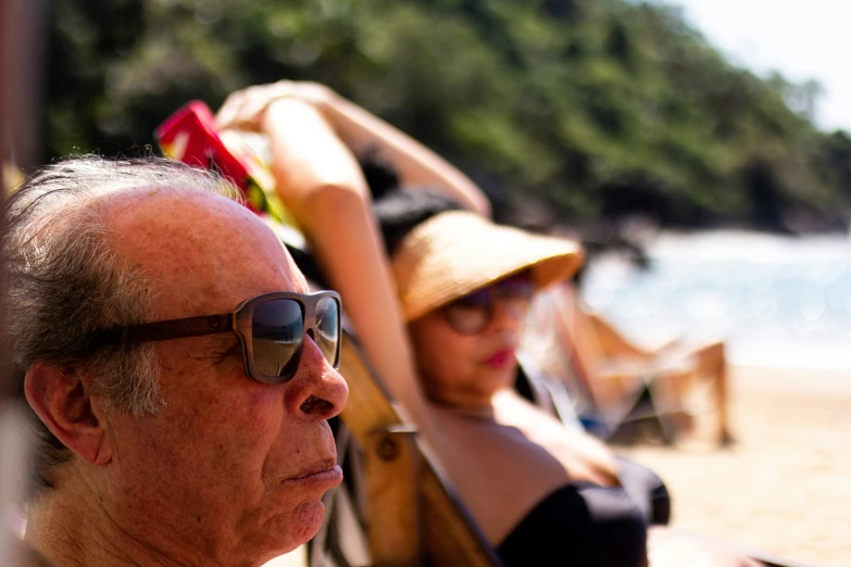 an older man and a young woman with hat, sun glasses and hats on sitting on a beach