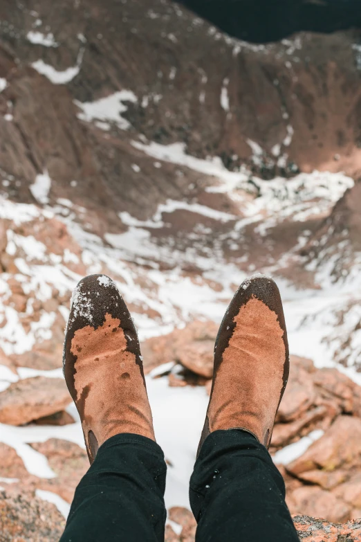 person in black pants and brown sneakers standing on rocks overlooking snow capped mountains