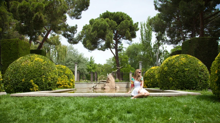 a woman sitting on a green grassy field near bushes and trees